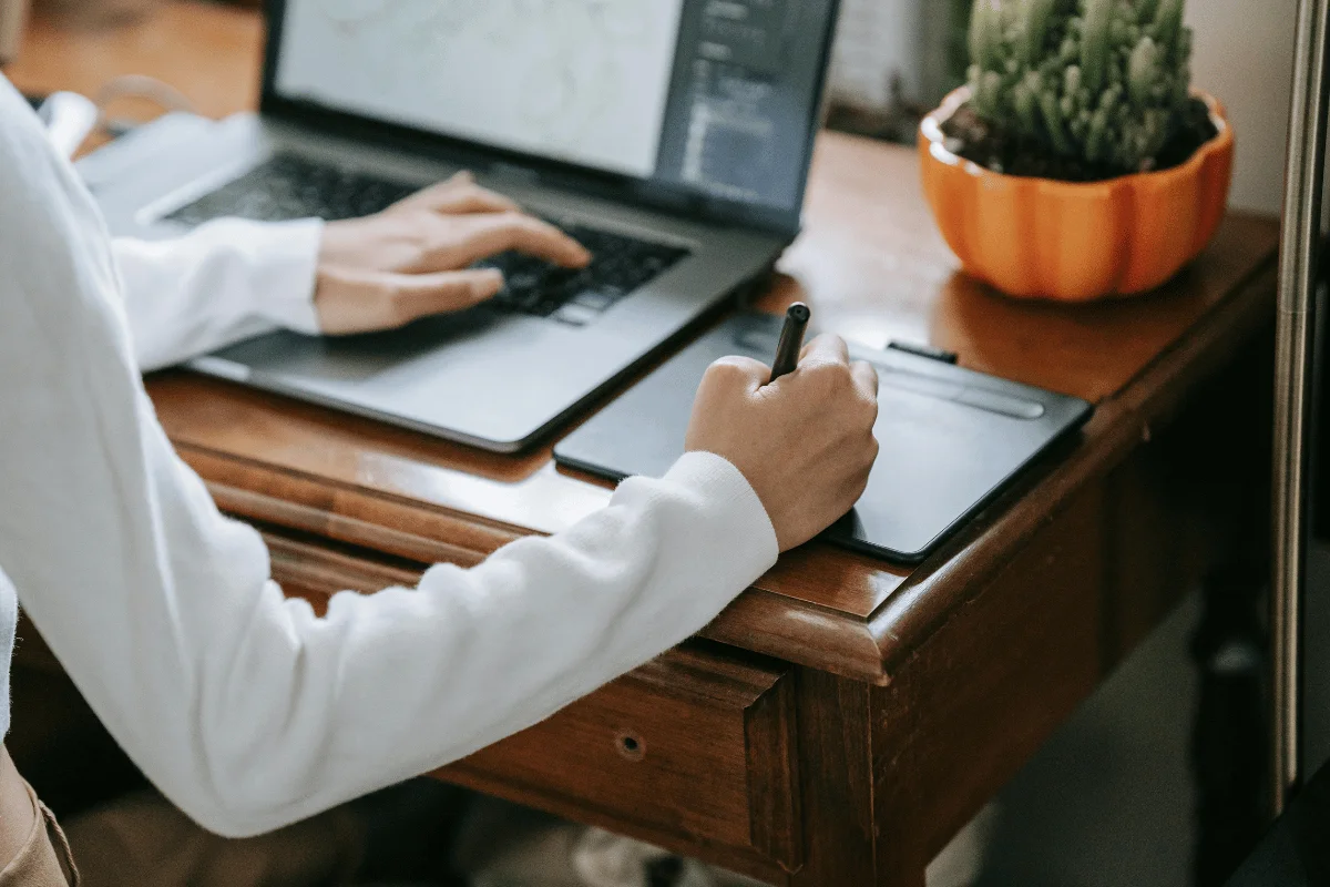 A person in a white sweater uses a stylus on a graphics tablet next to a laptop on a wooden desk. A small cactus in an orange pot adds a cozy touch.