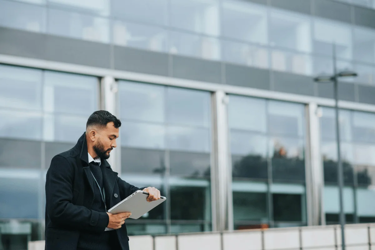 A man in a coat holding a laptop outdoors in front of a modern glass building.