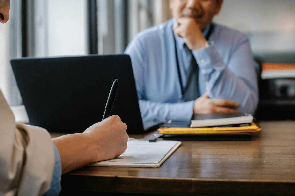 Close-up of business professionals in a meeting, with one person taking notes and the other sitting in front of a laptop.