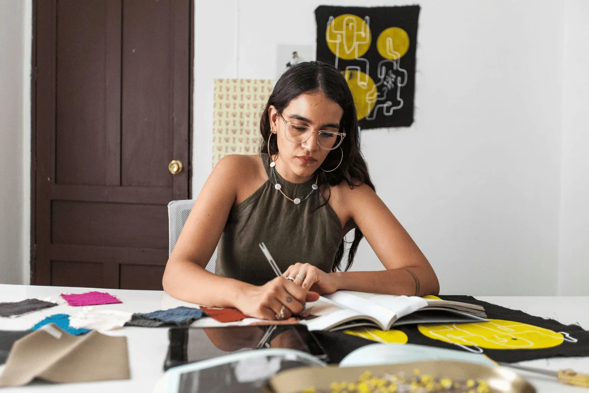 A young woman in a studio, wearing glasses, drawing in a notebook surrounded by colorful fabric swatches.