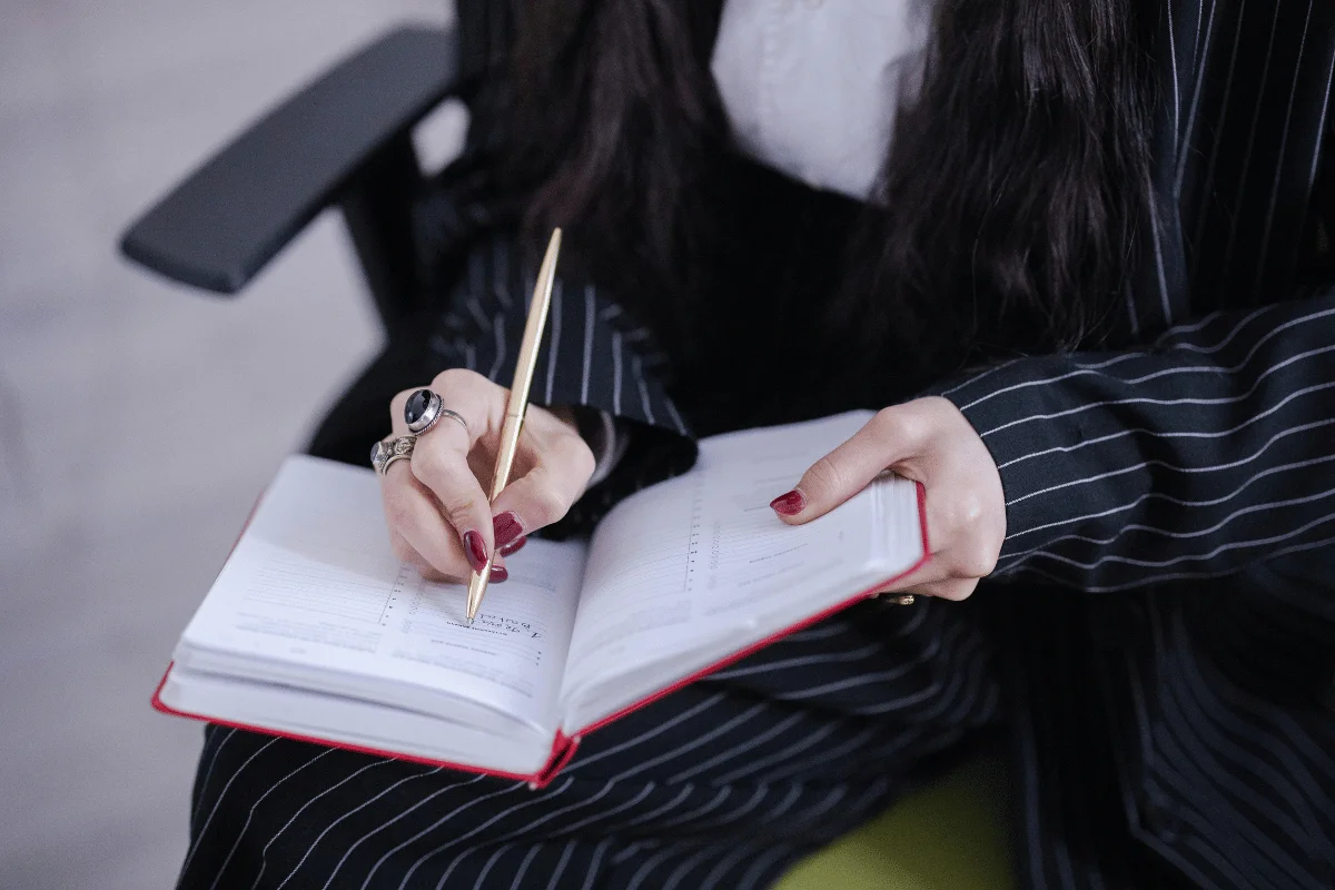 Close-up of a woman’s hands with red nails holding a notebook and pen.