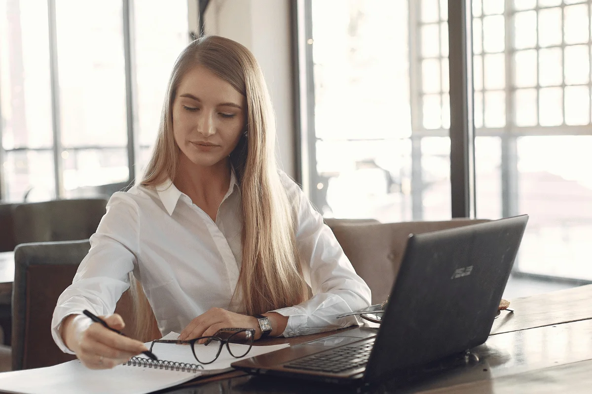 A woman with long hair in a white shirt is working at a desk, writing in a notebook beside a laptop. The setting is a bright, modern office.