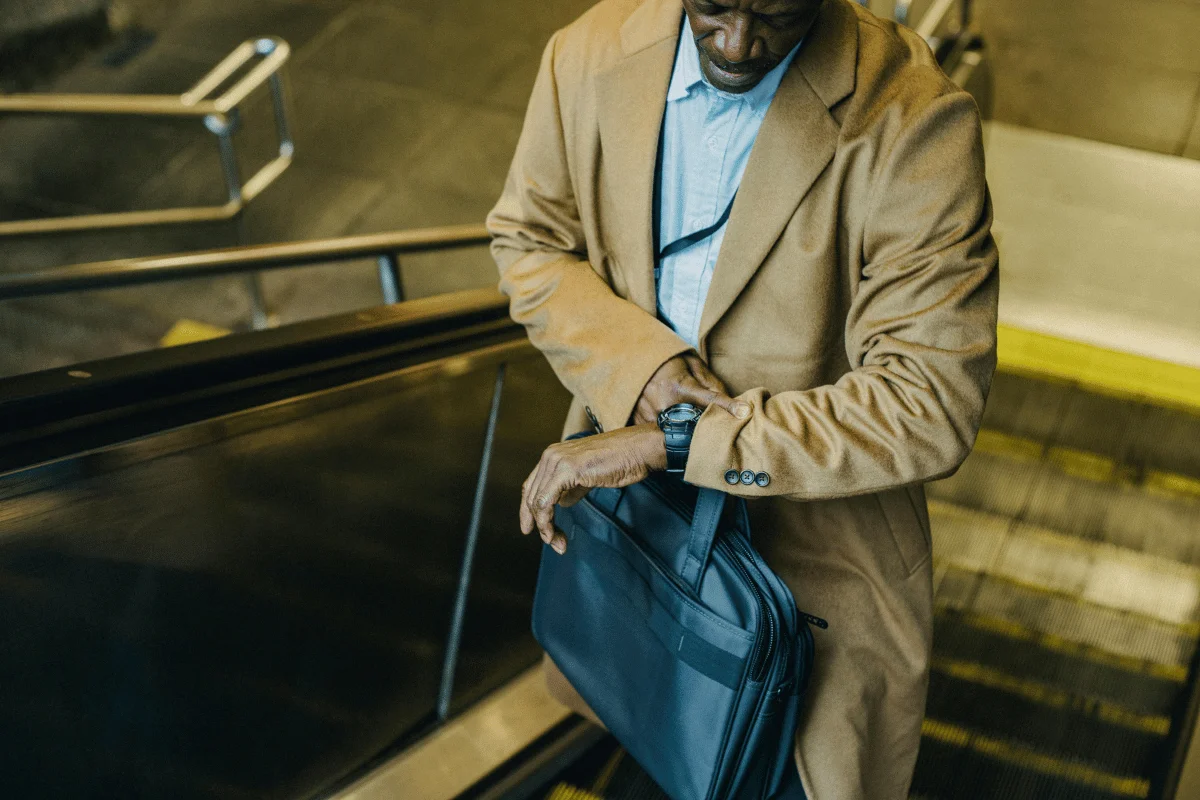 Man in a beige coat checking his watch while holding a laptop bag on an escalator.