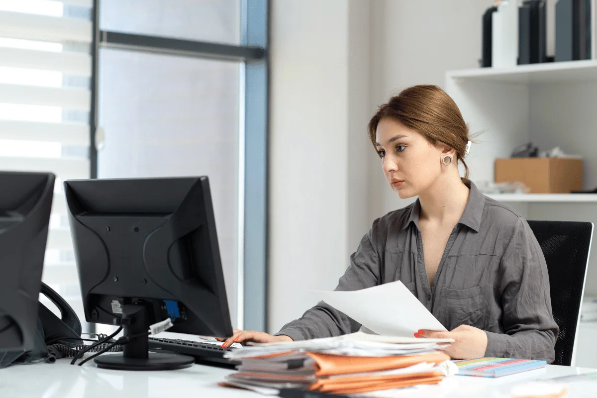 Woman working in an office, sitting at a desk with paperwork and two computer monitors.