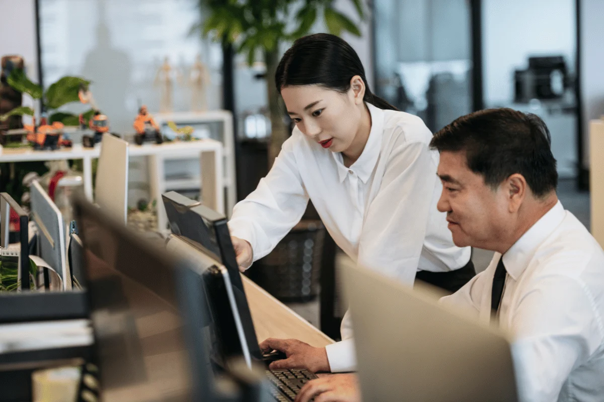 A young woman helping an older colleague with a computer task in a modern office.