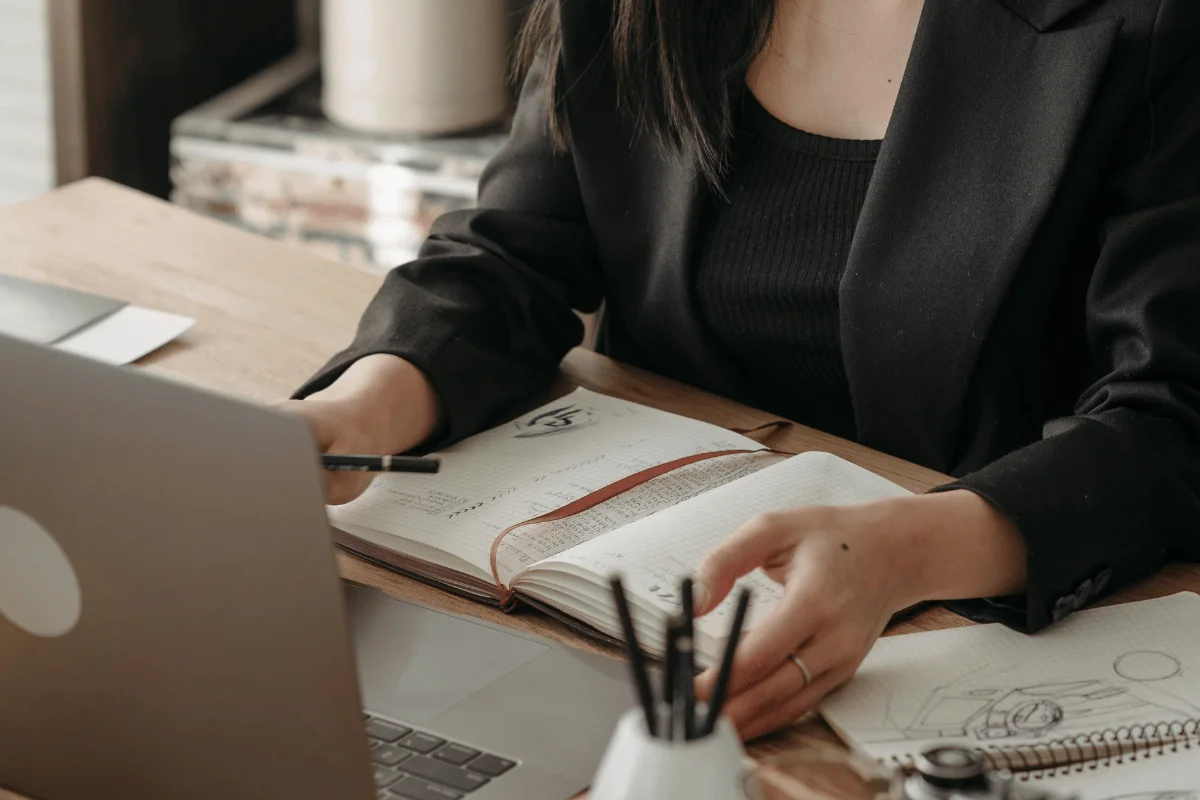A woman in a black blazer writes in a notebook while using a laptop at a desk.