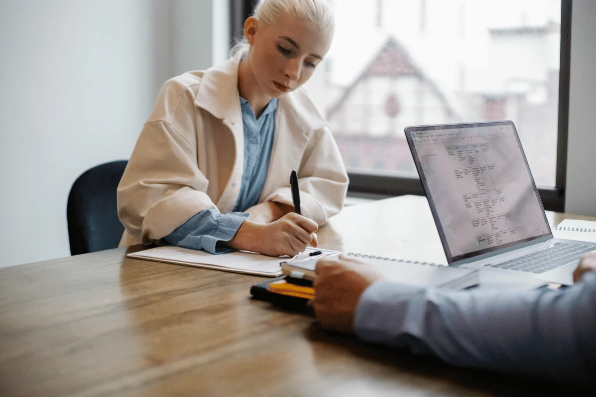 Two professionals having a meeting, with one writing notes while the other looks at a laptop screen with data.