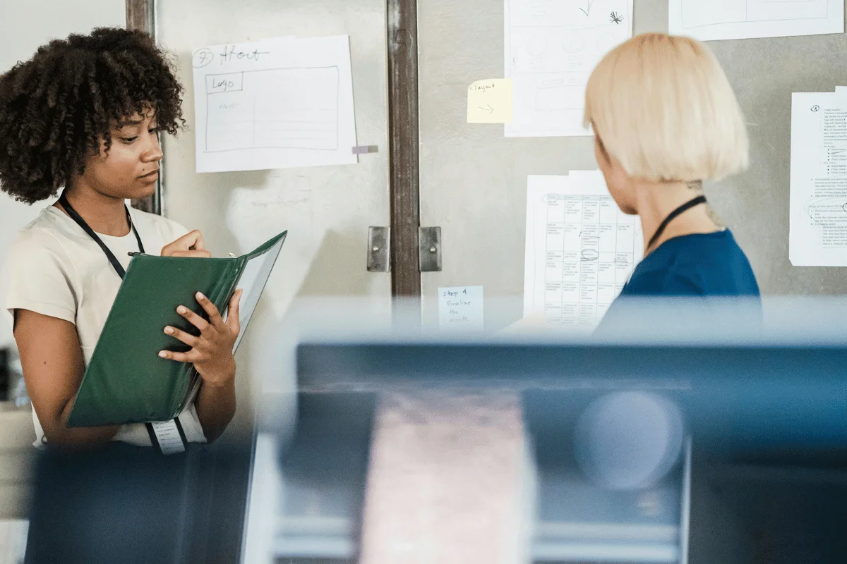 Woman in office holding a binder, listening attentively to a colleague in front of a wall with brainstorming notes and diagrams.