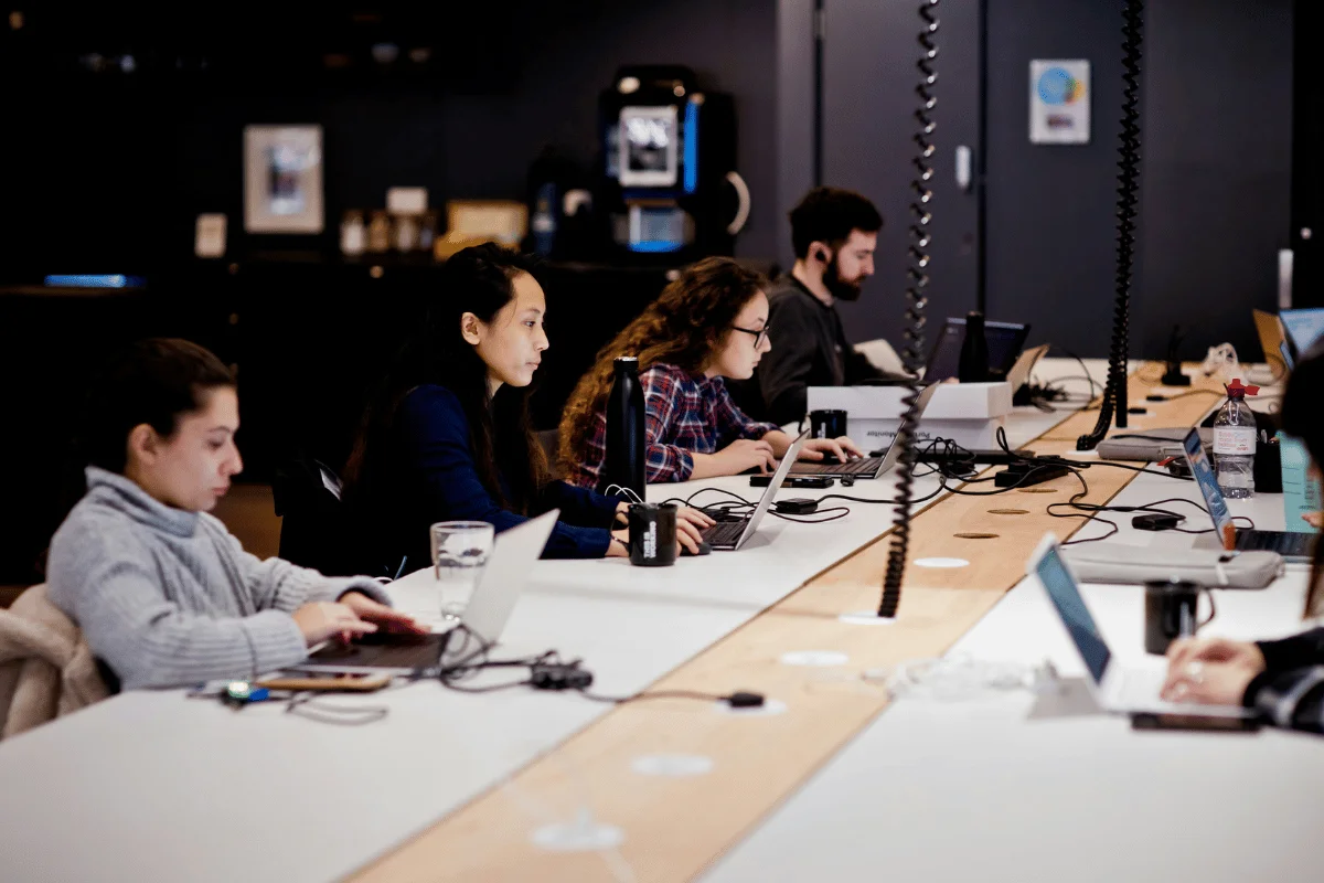 A group of people sits around a long table, focused on their laptops and devices in a modern workspace.