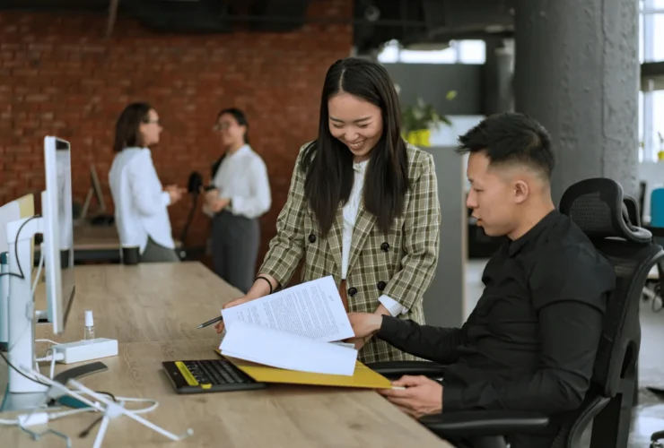 Two professionals discuss documents at a wooden desk, with computers and colleagues collaborating in a modern office setting.