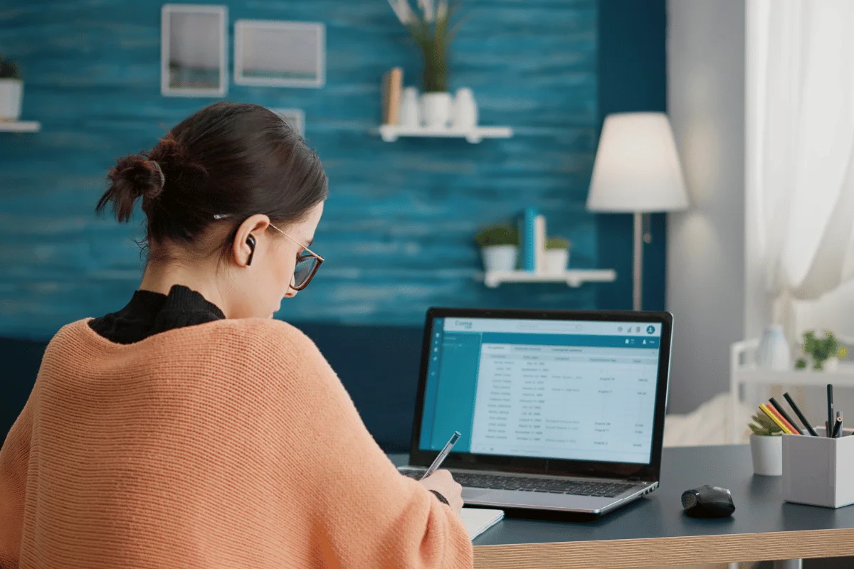 Woman working at a desk with a laptop and writing notes, surrounded by office supplies in a cozy workspace.