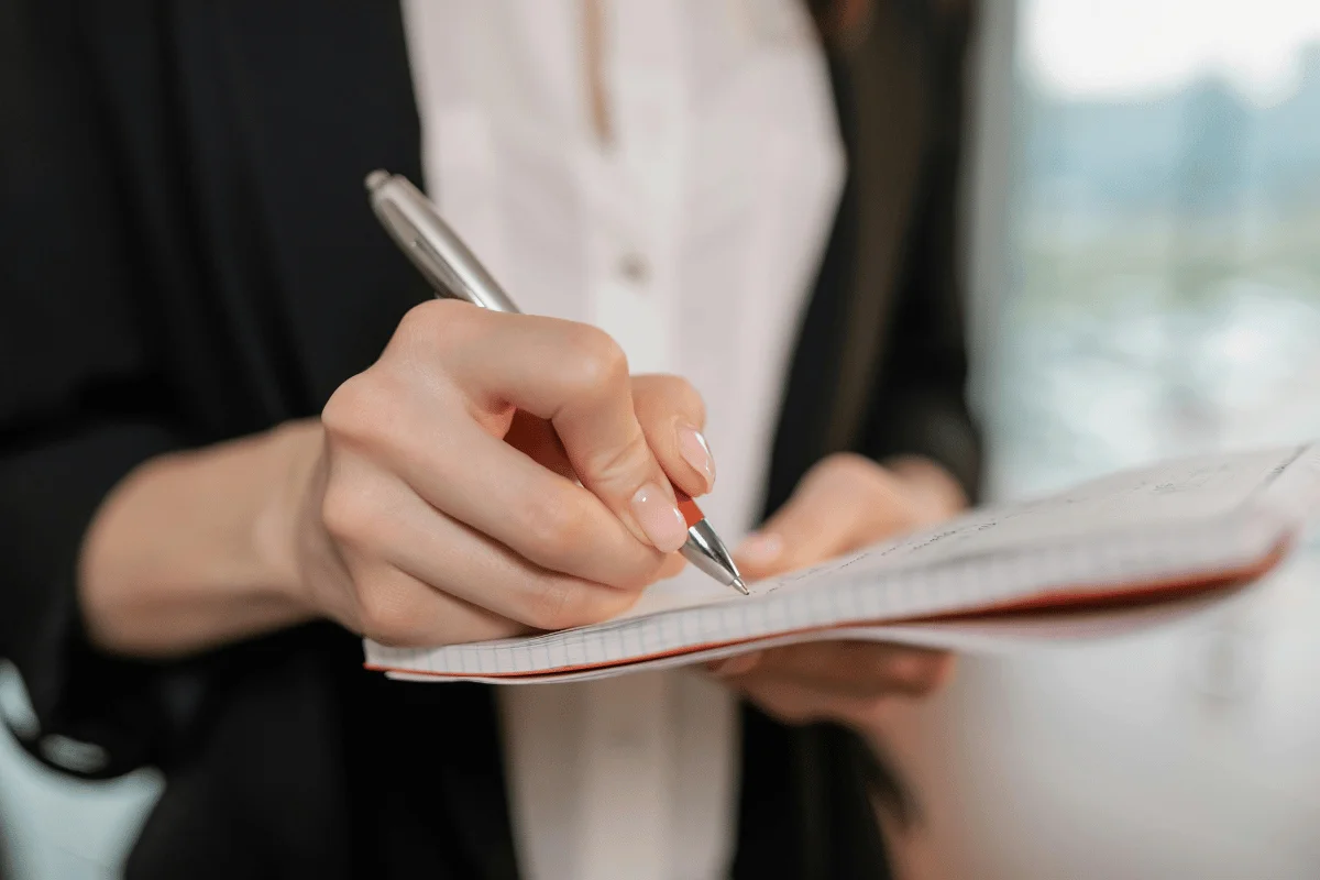 A close-up of a professional woman writing in a notebook with a silver pen.