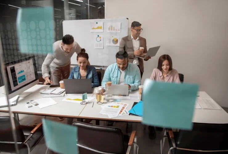 A diverse group collaborates in a modern conference room, analyzing data on laptops and charts, surrounded by coffee cups and papers.