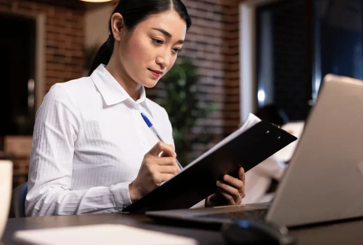 A person in a white shirt sits at a desk, holding a clipboard and pen, with a laptop and styled interior in the background.