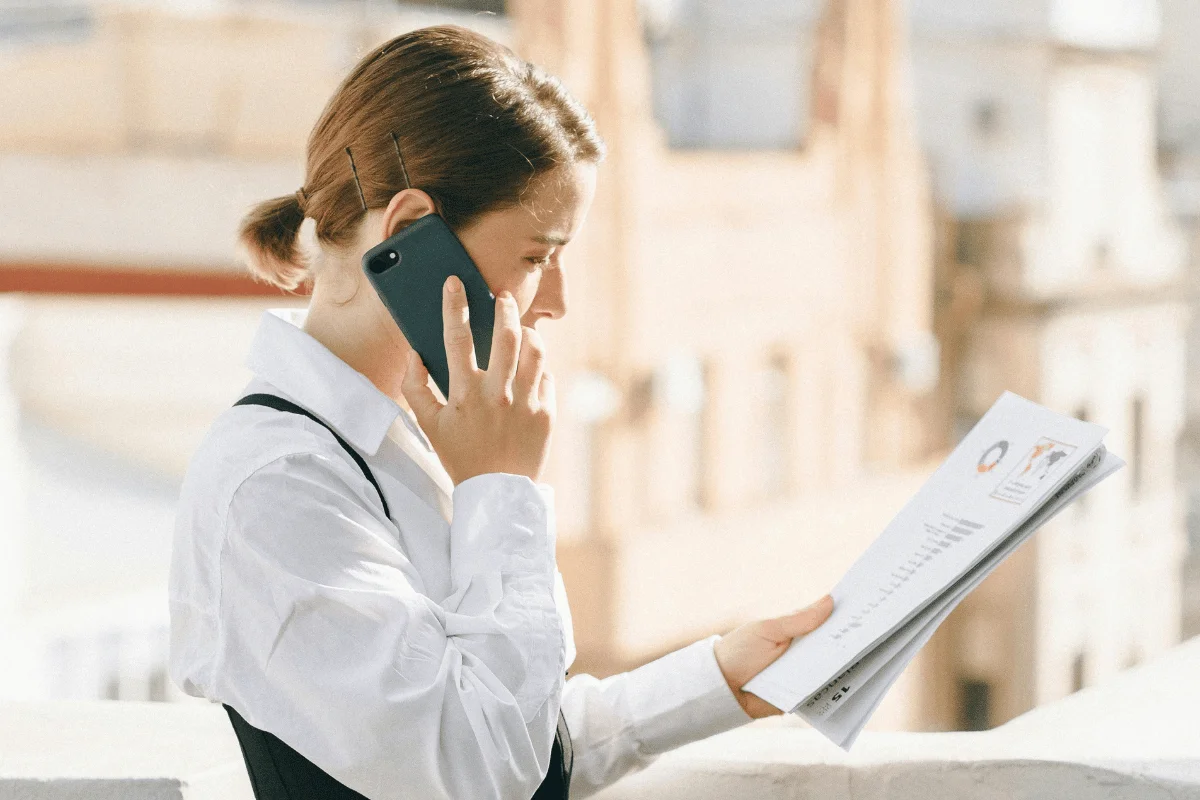 Businesswoman in a white blouse talking on the phone while reviewing documents outdoors.