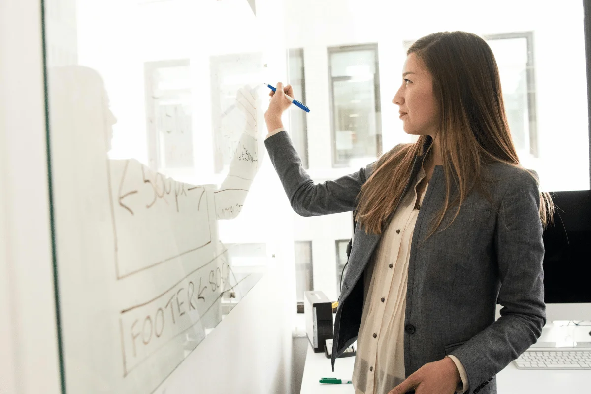 A woman in a business suit writes on a transparent whiteboard in a modern office setting.