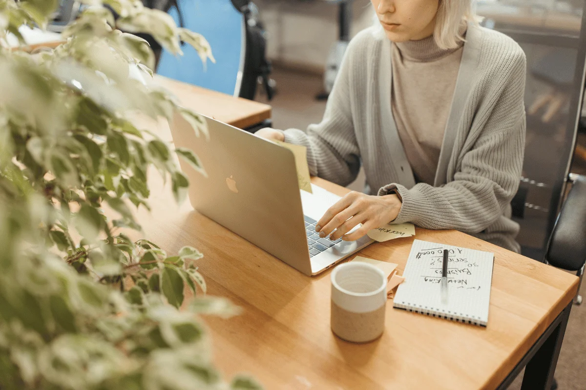 A young woman in a beige sweater typing on a laptop while taking notes in a notebook at a desk.