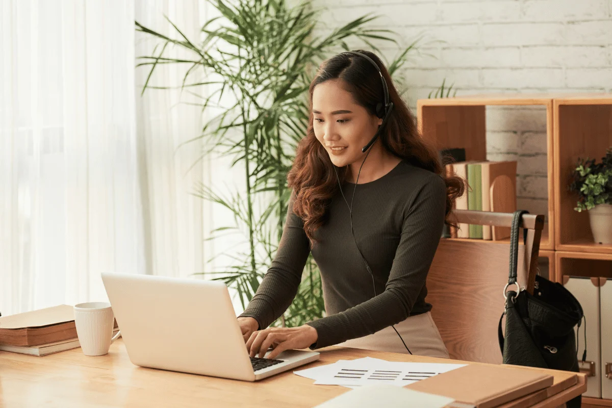 A woman with a headset typing on a laptop while working at a desk in a bright, airy room.