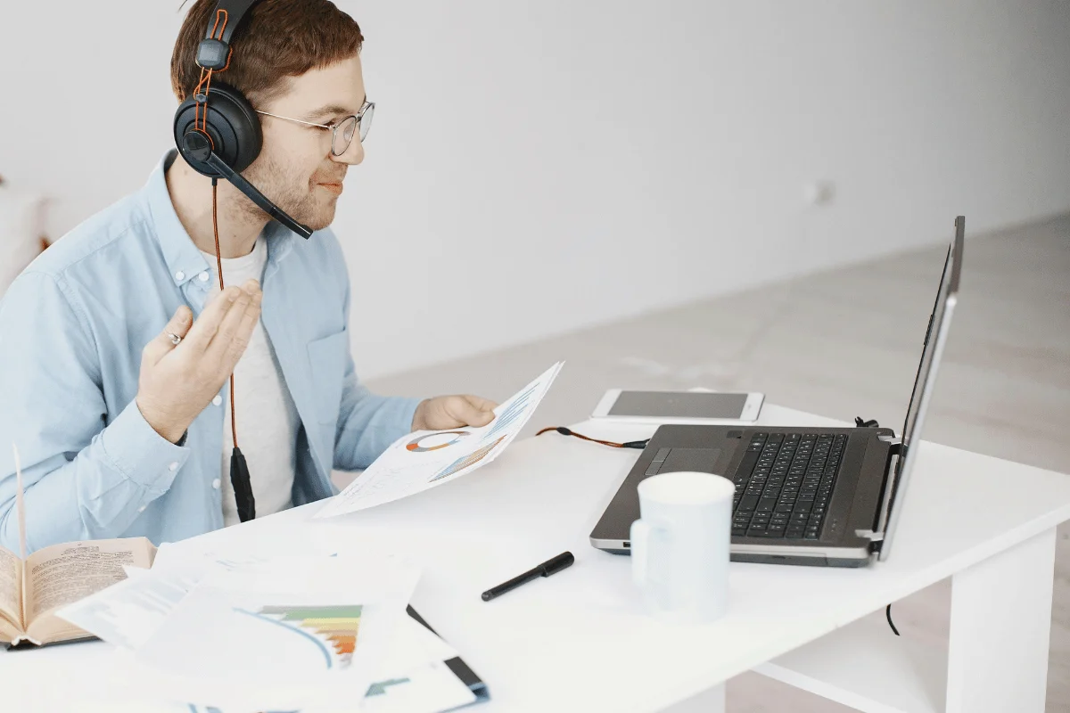 An individual wearing headphones gestures while reviewing documents at a desk with a laptop, tablet, and a cup.