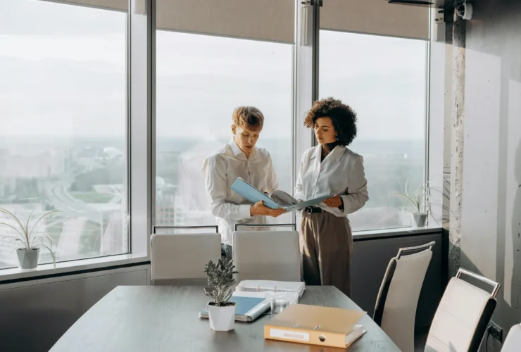 Two professionals engaged in discussion over documents, with a cityscape visible through large windows behind them. A modern conference room setting.