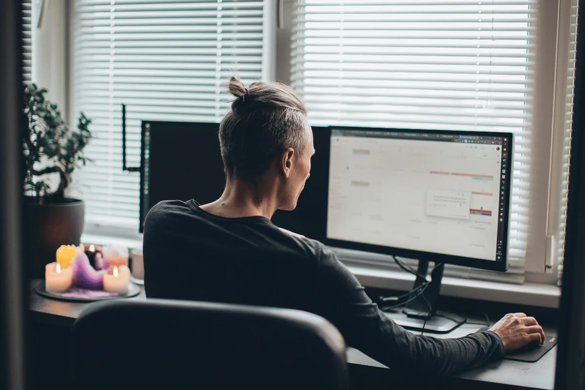 Freelancer using a dual-screen setup in a cozy office with natural light from blinds.