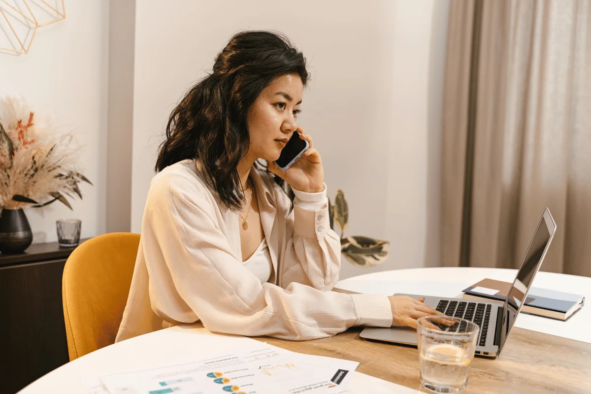 A woman talks on the phone while working on a laptop at a wooden table, with documents and a glass of water beside her.