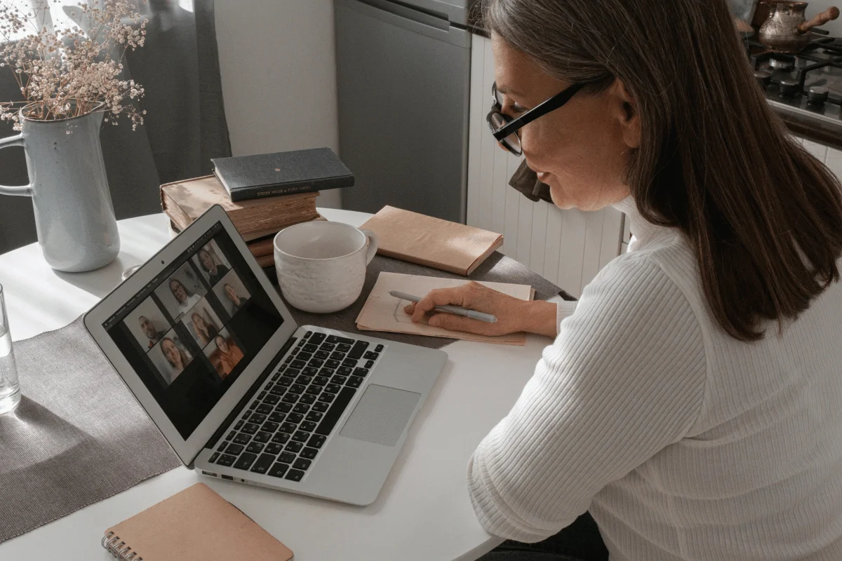 A person in a white sweater participates in a video call on a laptop at a table, surrounded by books, a mug, and a notebook.