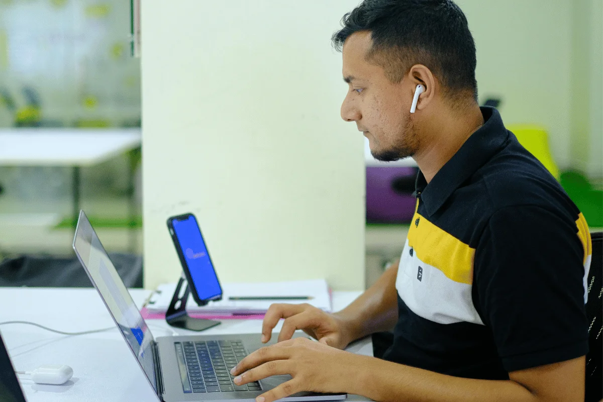 A person wearing AirPods uses a laptop while seated at a desk with a mobile phone and a notebook in an office setting.