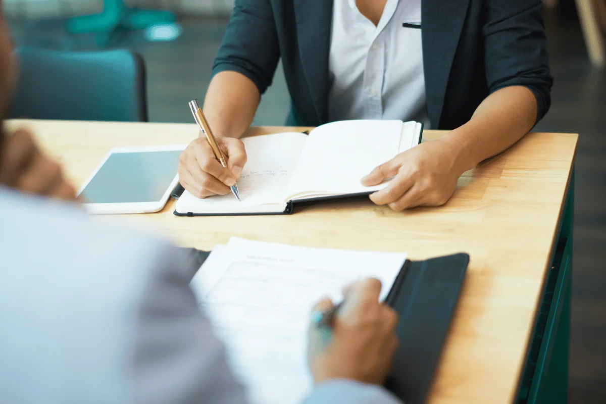 A close-up of two individuals in a business meeting; one writes in a notebook while the other gestures with paperwork in front.