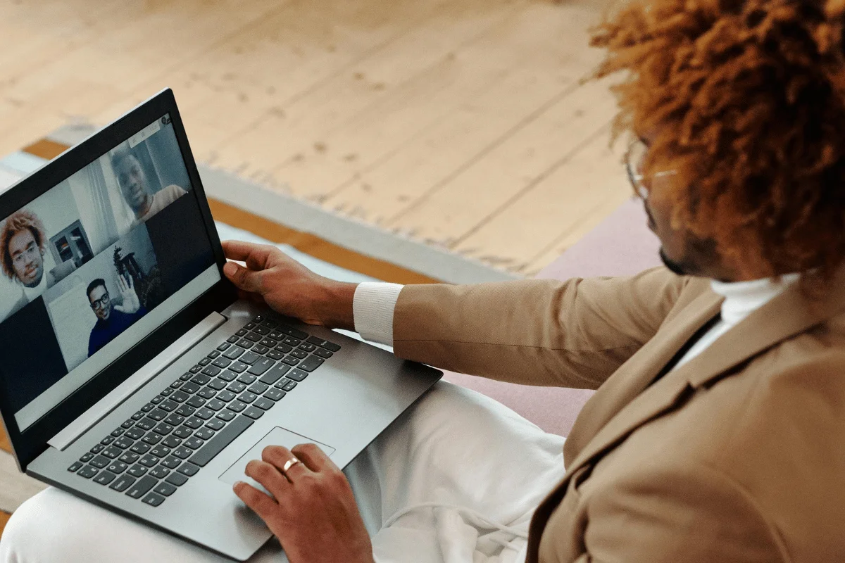 A person in a beige suit sits on a sofa, engaged in a video call on a laptop, with various remote participants displayed on screen.
