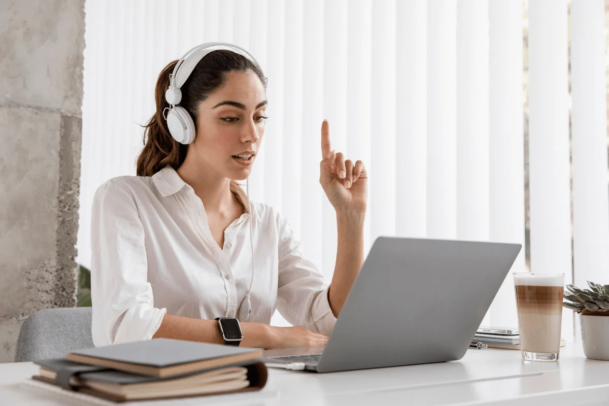 A person in a white shirt and headphones gestures thoughtfully while working on a laptop, surrounded by books and a coffee cup.