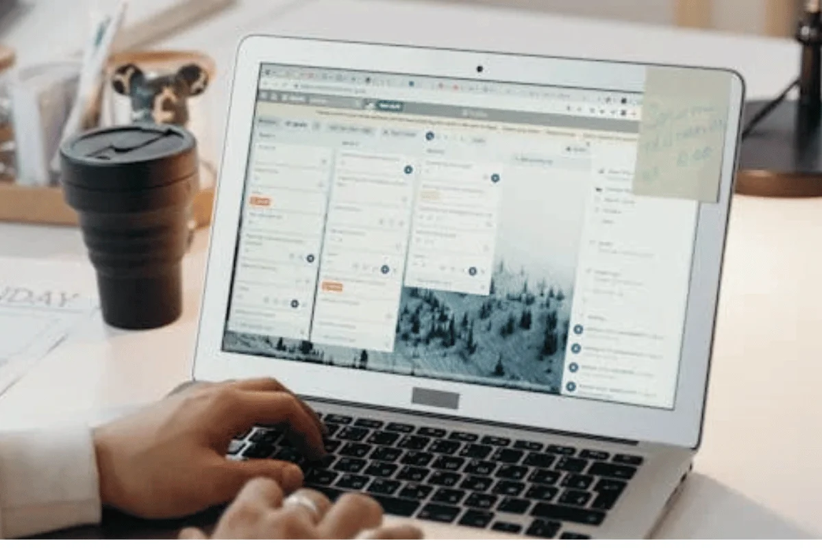 An individual's hand on a laptop displaying a productivity app, with a coffee cup and workspace items in the background.