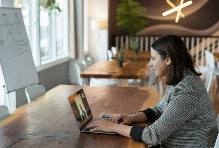A woman sits at a wooden table, engaged in a video call on her laptop in a modern workspace with plants and a whiteboard in the background.