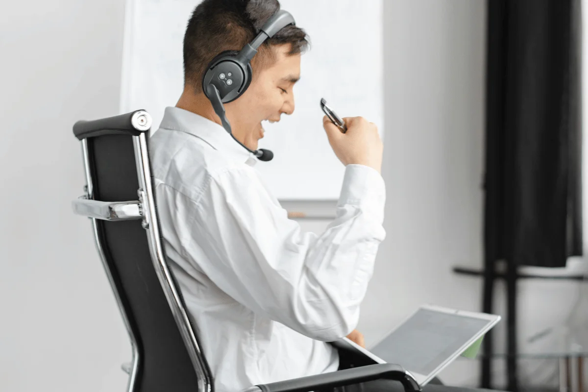A person in a white shirt sits in a black ergonomic chair, wearing a headset and holding a pen, with a laptop nearby.