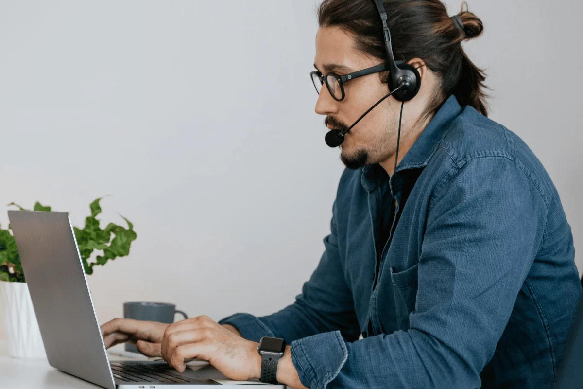 A person wearing headphones types on a laptop at a desk adorned with a potted plant and a coffee mug, suggesting a focused work environment.