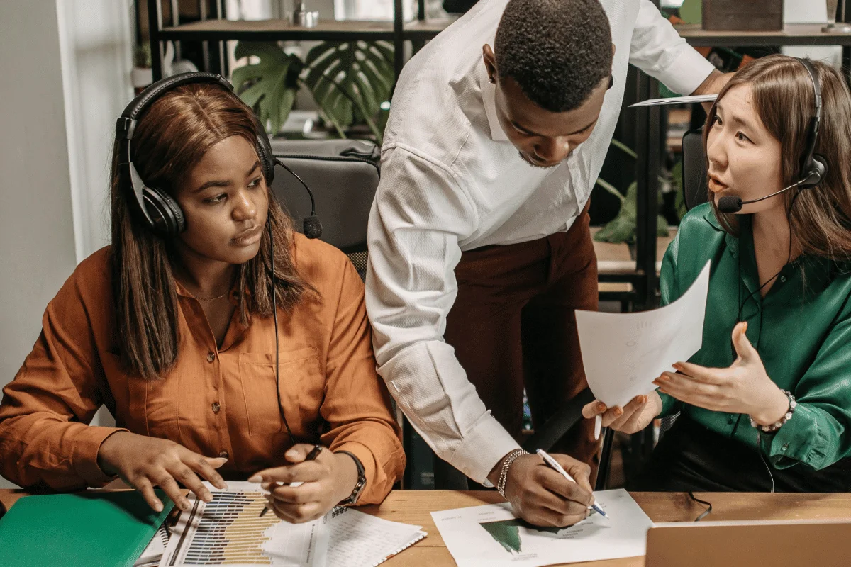 A diverse group of professionals collaborates at a desk, discussing documents and analyzing data in a modern, green office space.