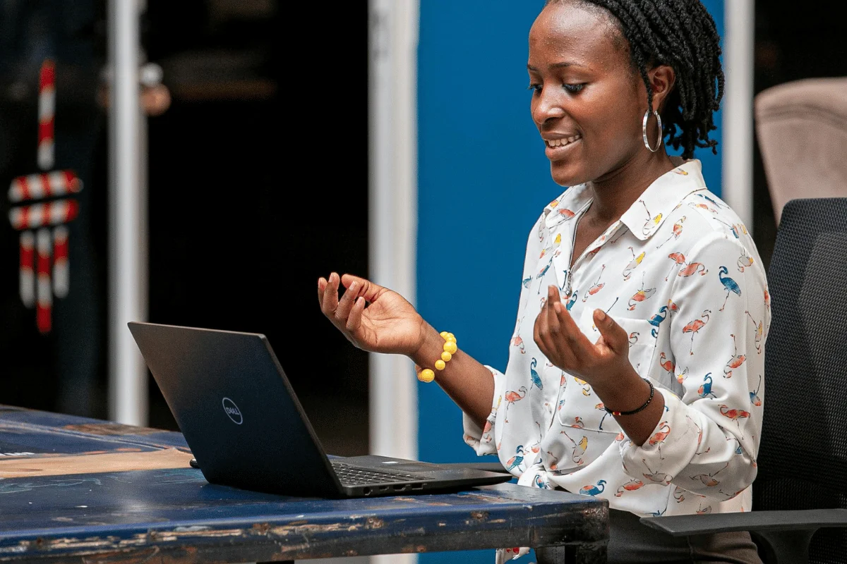 A person gestures expressively while sitting at a table with a laptop, wearing a white shirt adorned with colorful flamingo patterns.