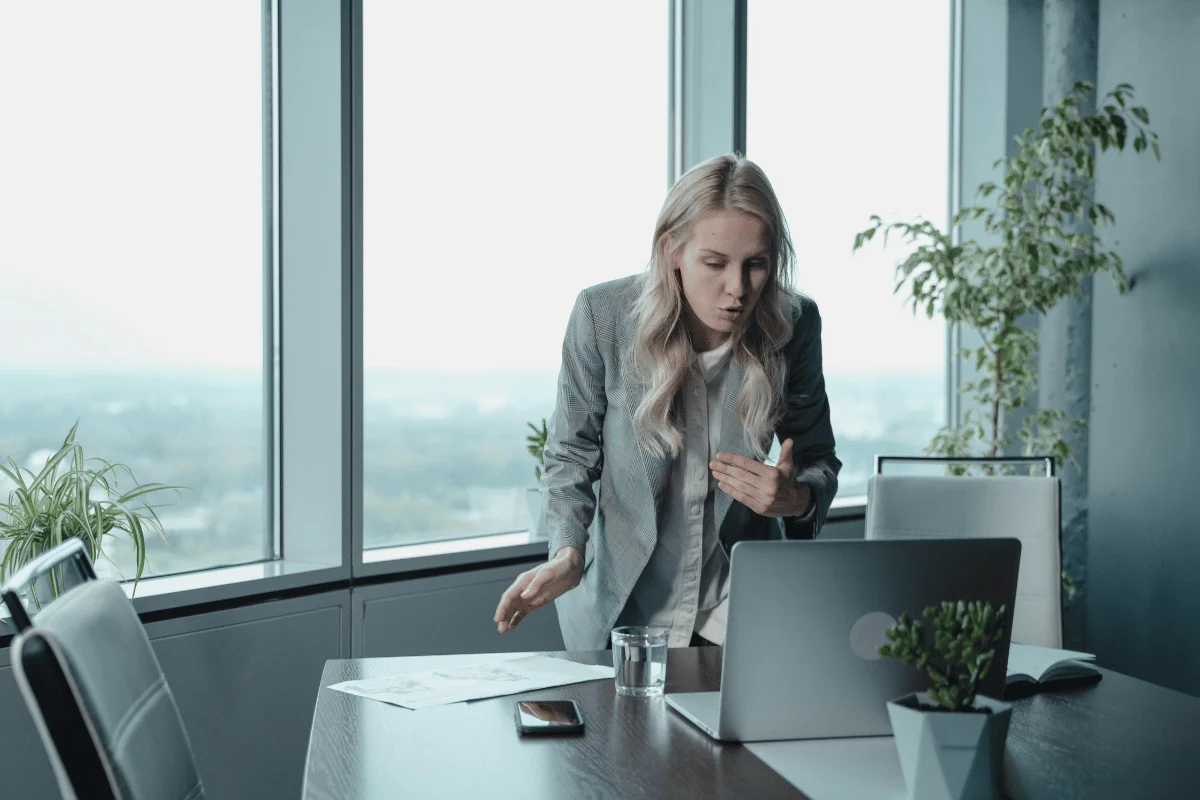 A woman in a business suit gestures while standing near a desk with a laptop, plants, and documents in a modern office with large windows.