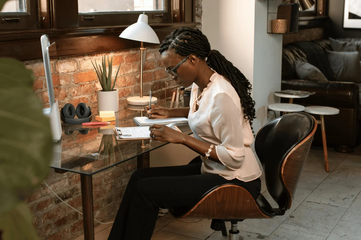 A focused woman in a pale shirt sits at a glass desk, writing notes surrounded by a cozy, modern workspace with brick walls and plants.