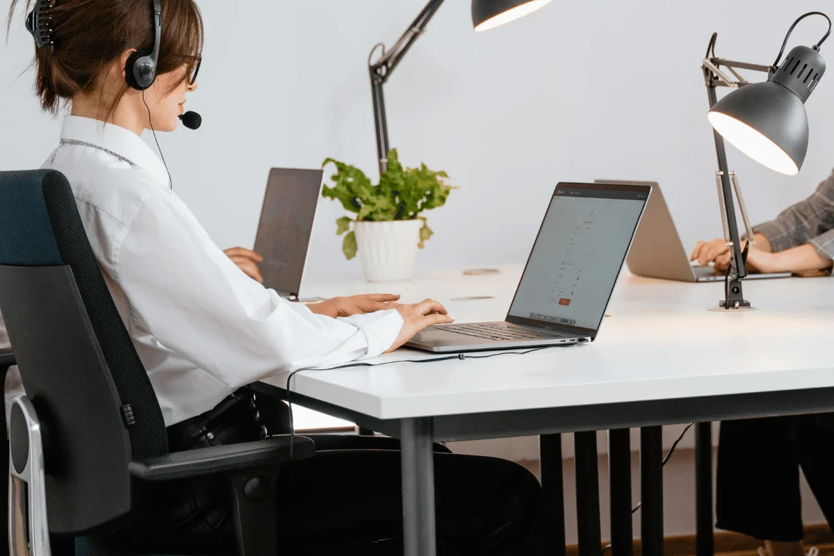 A person wearing a headset is typing on a laptop at a bright workspace with potted plants and workstations.