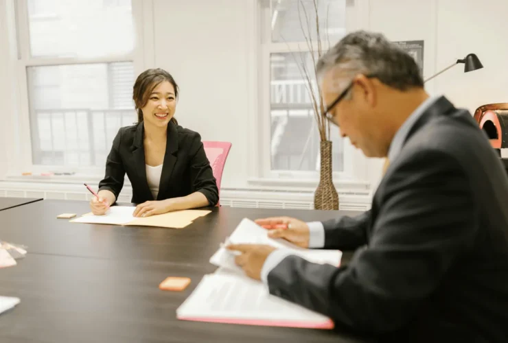 A woman in a blazer writes on paper at a conference table while a man in a suit speaks, surrounded by bright office decor.