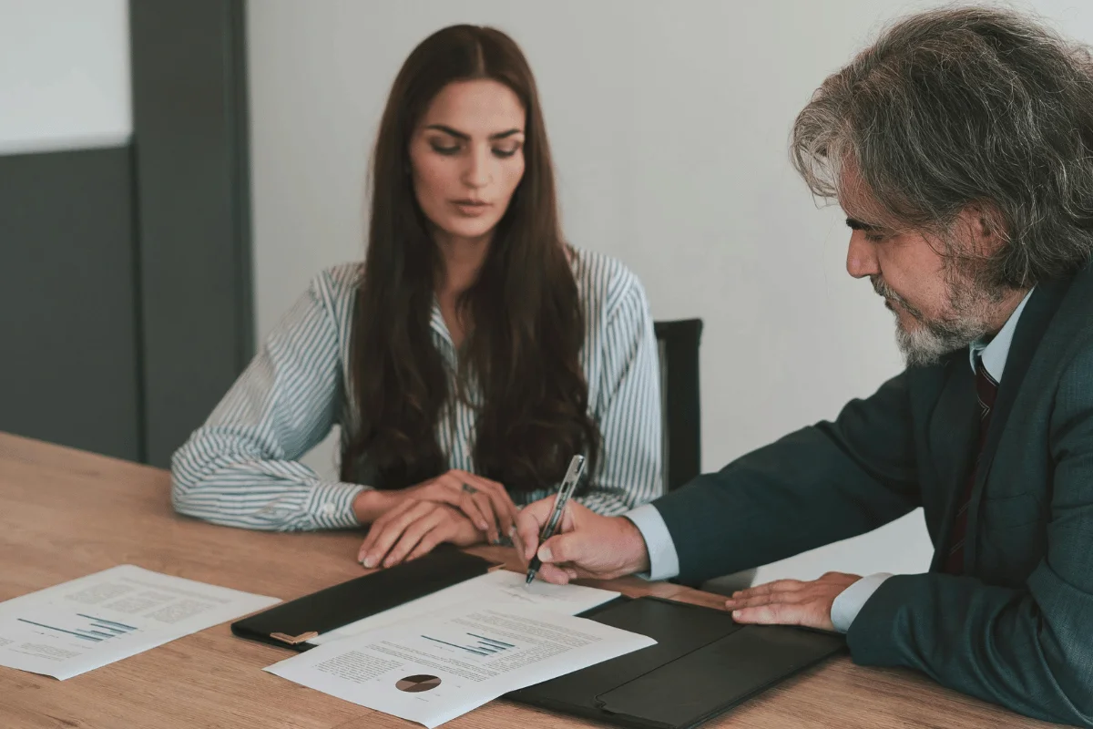 Serious individuals in a meeting scene with two people signing documents on a wooden table, surrounded by papers and a portfolio.