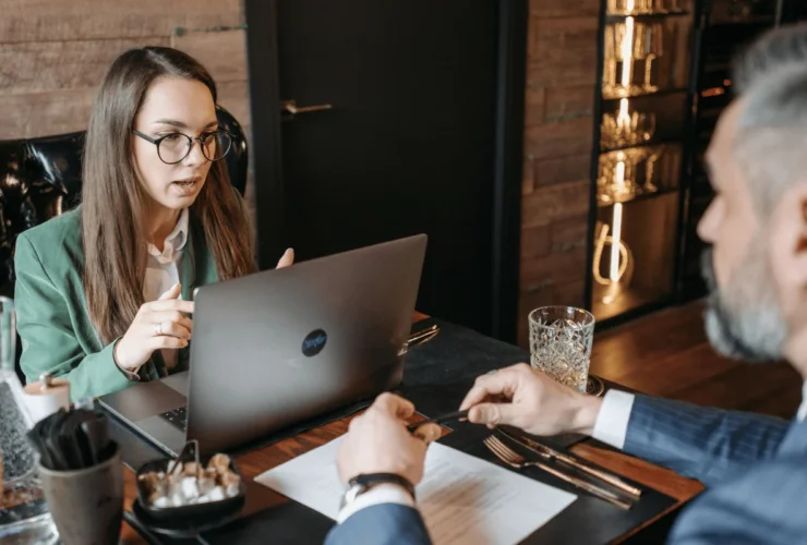 A woman in a green blazer engages in conversation at a desk with a laptop, while a man gestures with a pen across from her.