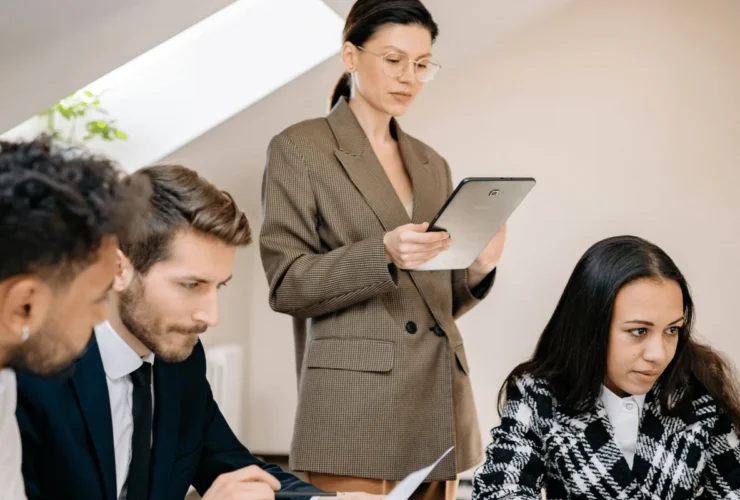 A business meeting in progress with a presenter holding a tablet while seated individuals engage with notes and documents.