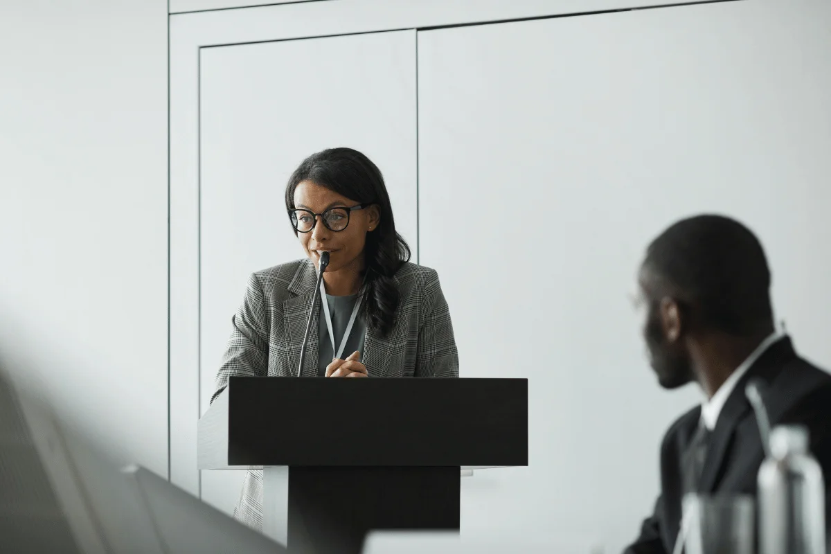 A woman speaking at a podium with a microphone in front of an audience in a conference room.