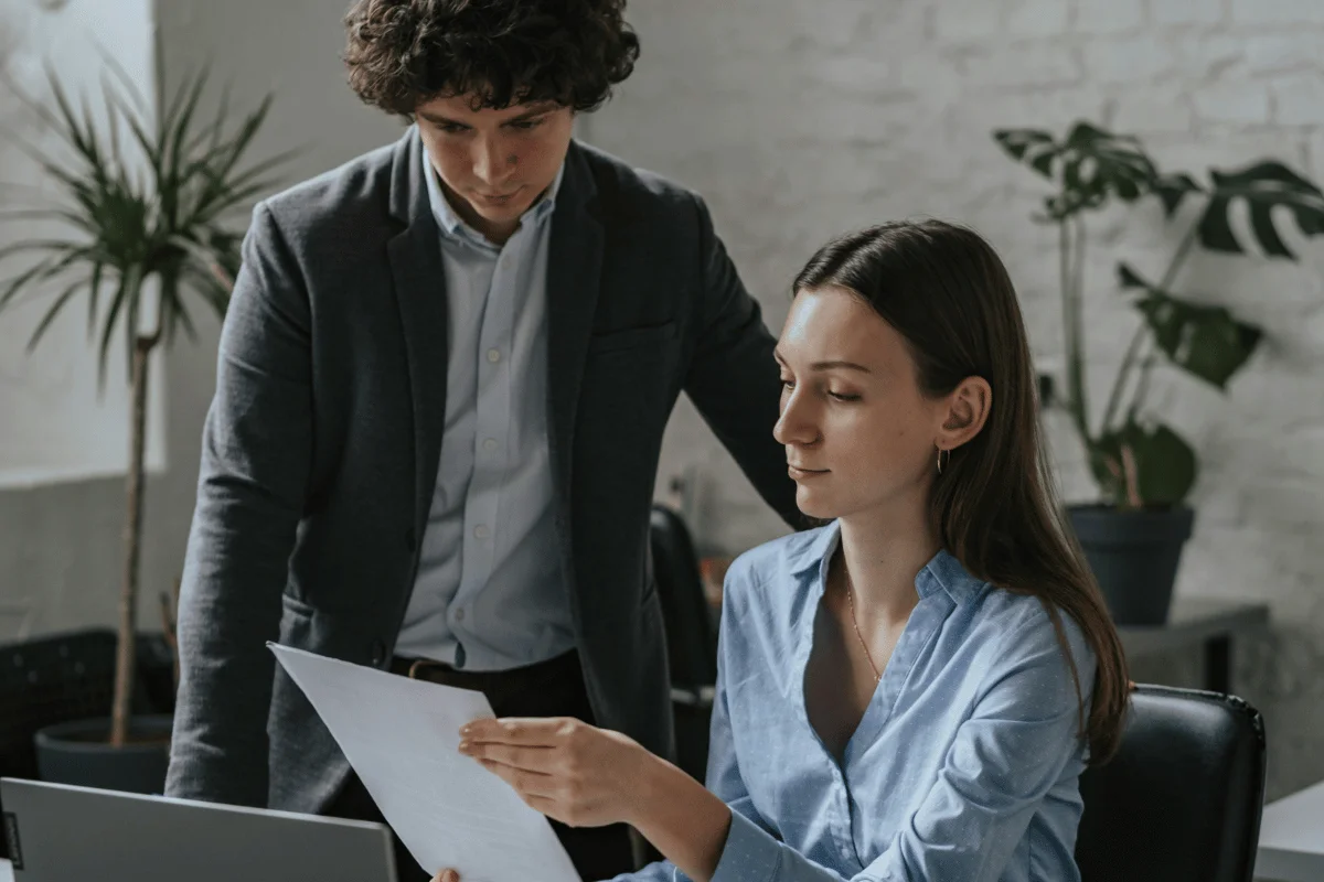 A woman in a blue shirt sits at a desk, holding a document, while a man in a dark blazer stands beside her, both appearing focused. Indoor plants decorate the background.