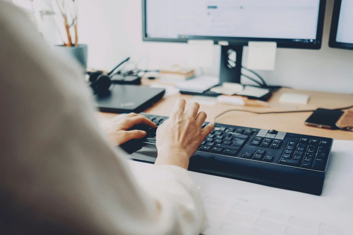 A person types on a black keyboard at a desk with dual monitors. 