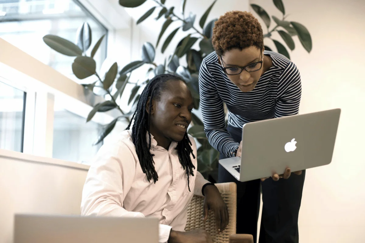 Two people collaborate in a well-lit office, focusing intently on a laptop. Lush greenery adds a calm, engaging backdrop to their teamwork.