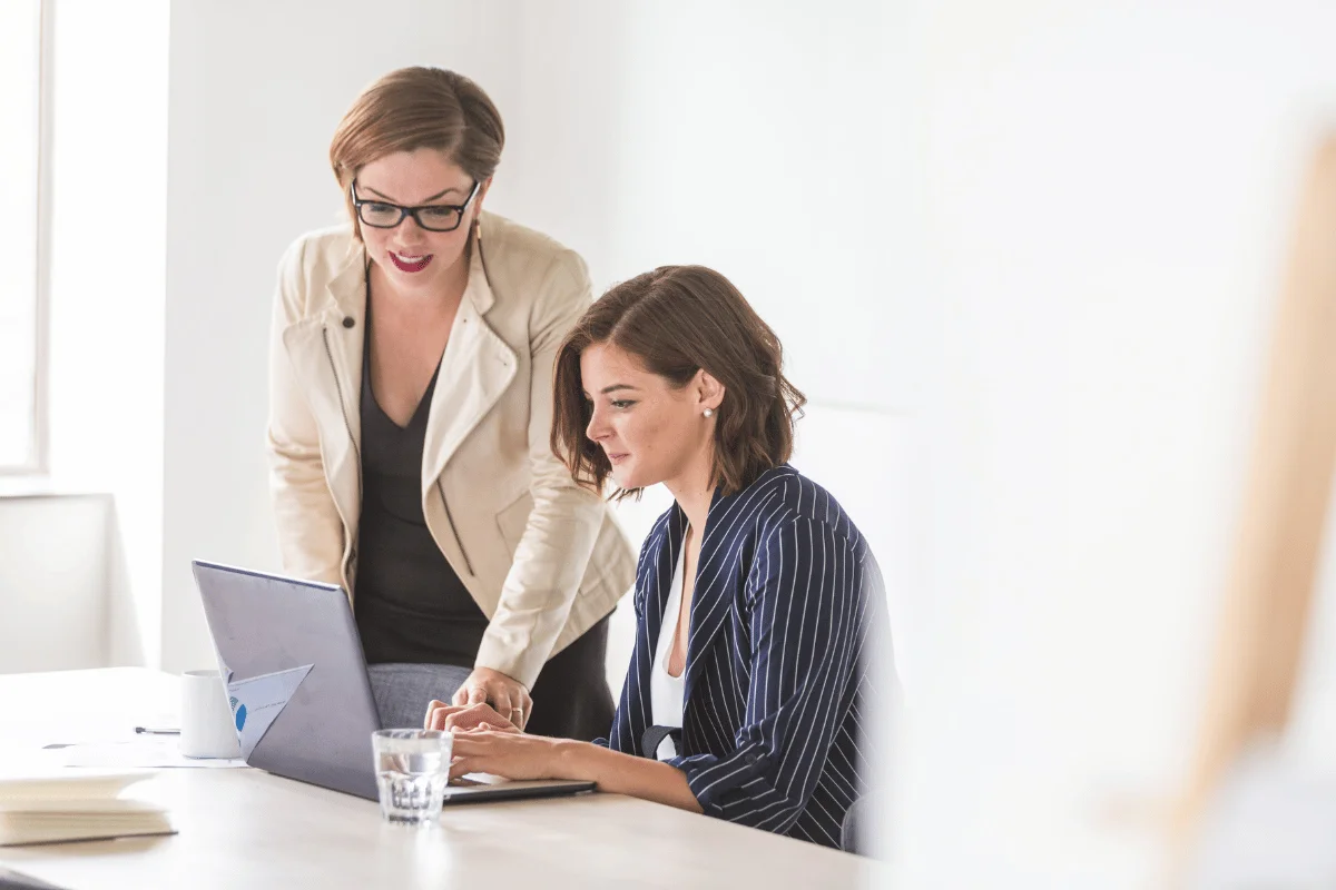 Two women collaborate at a desk with a laptop. One is seated typing, while the other stands, leaning over and giving suggestions. Bright and focused.