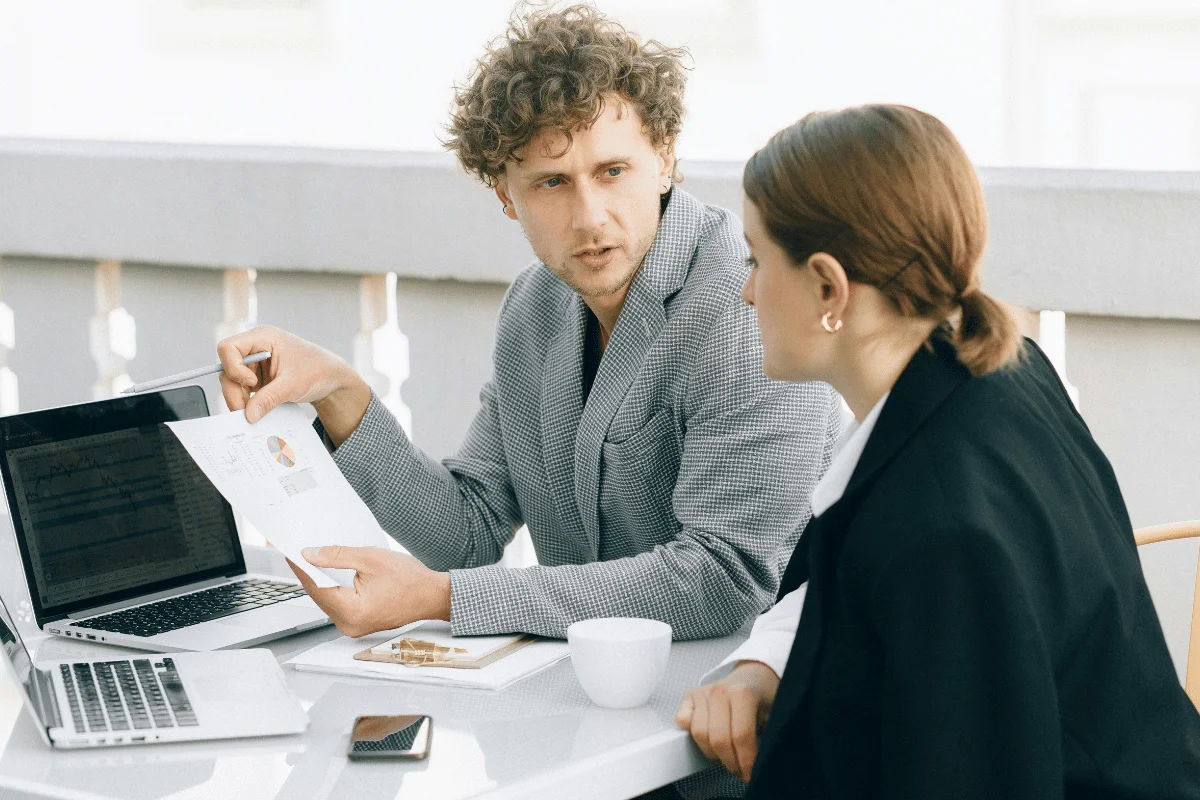 A curly-haired man in a checkered blazer discusses a document with a woman in a black coat. They're seated at an outdoor table with laptops, looking focused.